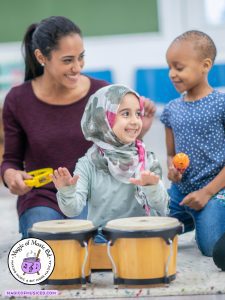 Smiling elementary music teacher and two young girls playing classroom instruments together, including bongos, a tambourine, and a maraca.