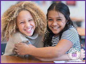 Two smiling elementary school girls hugging at a desk, radiating joy and friendship. This image represents the Friendship & Unity choir concert theme, perfect for celebrating connection and togetherness through music.