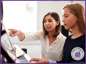 A music teacher in a white blouse gives encouraging feedback to a young piano student, pointing at the sheet music to highlight their progress. The teacher’s supportive approach helps build confidence and motivation in the music classroom. The Magic of Music Ed logo in the corner reinforces the theme of positive feedback in music class.