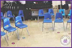 A music classroom setup with blue chairs arranged in a semicircle, each placed over colorful numbered spot markers on the floor. The background features instrument cases and percussion equipment.