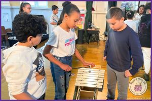 Three elementary students playing a metallophone in a general music classroom, exploring instruments that support a well-rounded music education on a classroom budget.