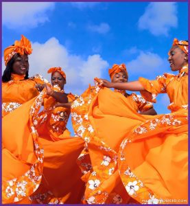Four women dressed in bright orange traditional Caribbean attire performing a joyful cultural dance under a blue sky, representing the spirit of Caribbean music and movement.