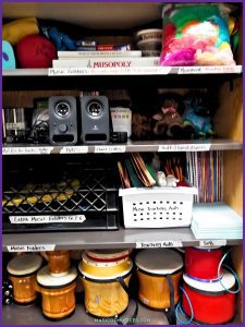Shelf filled with colorful hand drums, a violin, speakers, and stuffed animals for general music, showcasing affordable supplies for elementary music teachers managing a classroom budget.