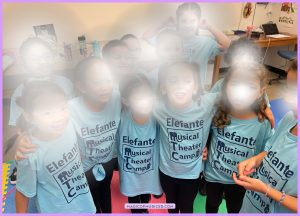 Elementary students in “Elefante Musical Theater Camp” shirts gathered in a classroom, showing music education benefits like friendship, teamwork, and confidence.