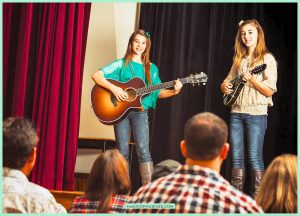Two students play guitar and mandolin on a school stage before an audience, illustrating music education and its benefits for confidence and collaboration.
