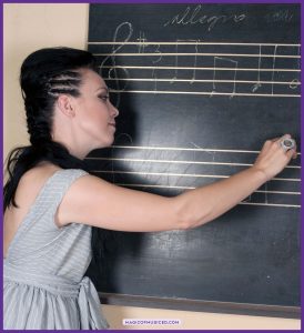 Music teacher writes a musical excerpt on a chalkboard staff with the tempo marking “allegro,” modeling notation skills for music performance assessment.